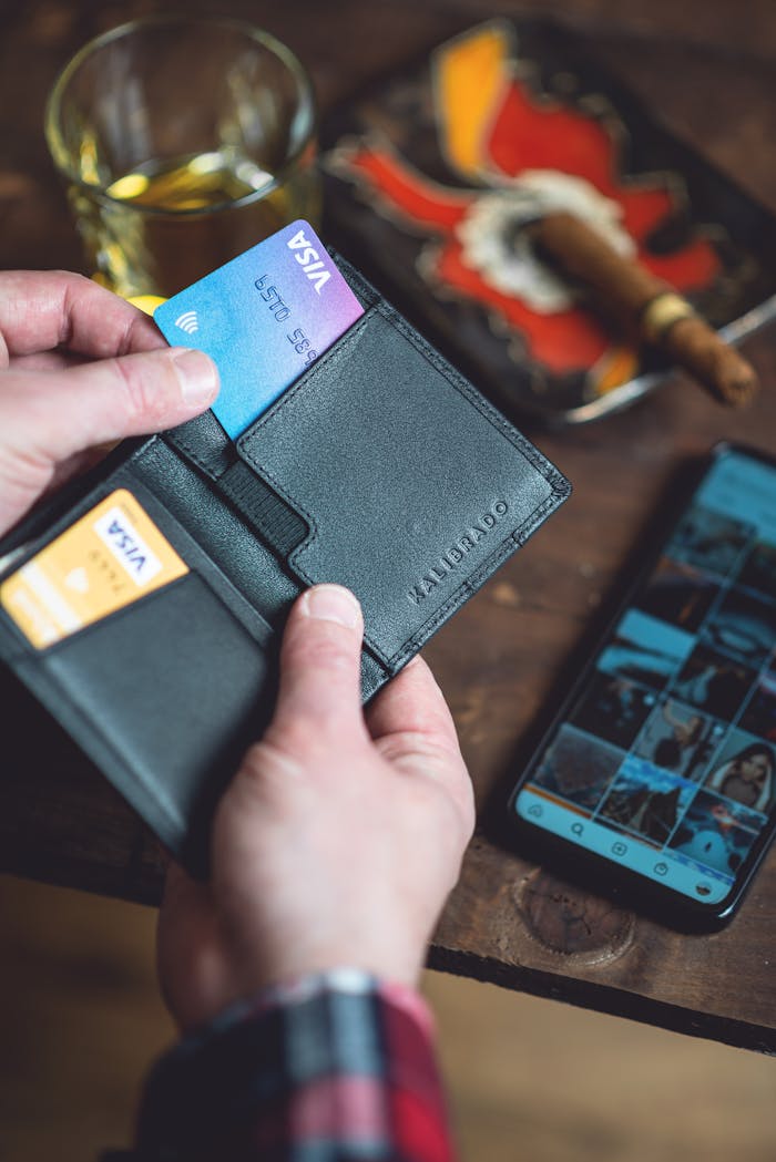 creative-01 Hands holding a sleek black wallet containing credit cards next to a smartphone on a wooden table.